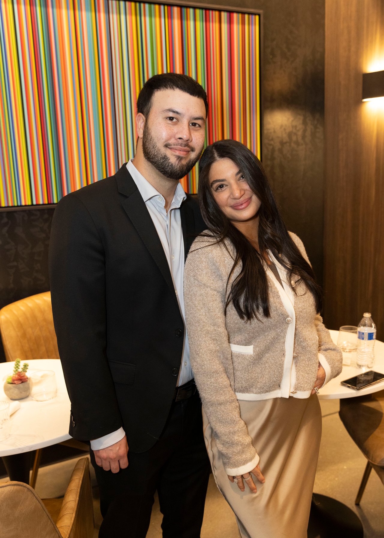 A man in a dark suit and a woman in a light cardigan pose together in front of colorful striped artwork.