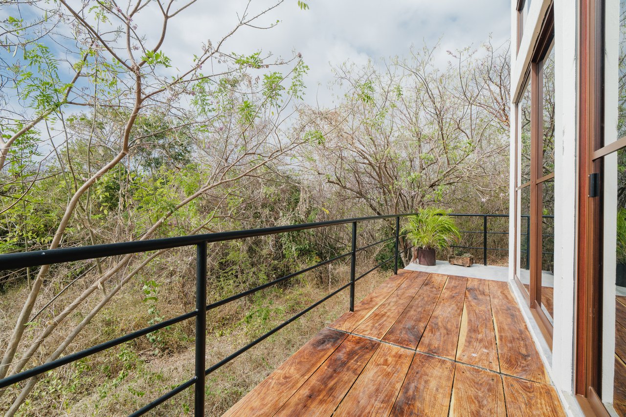 Wooden deck with black railing beside large sliding glass doors, overlooking dense trees.