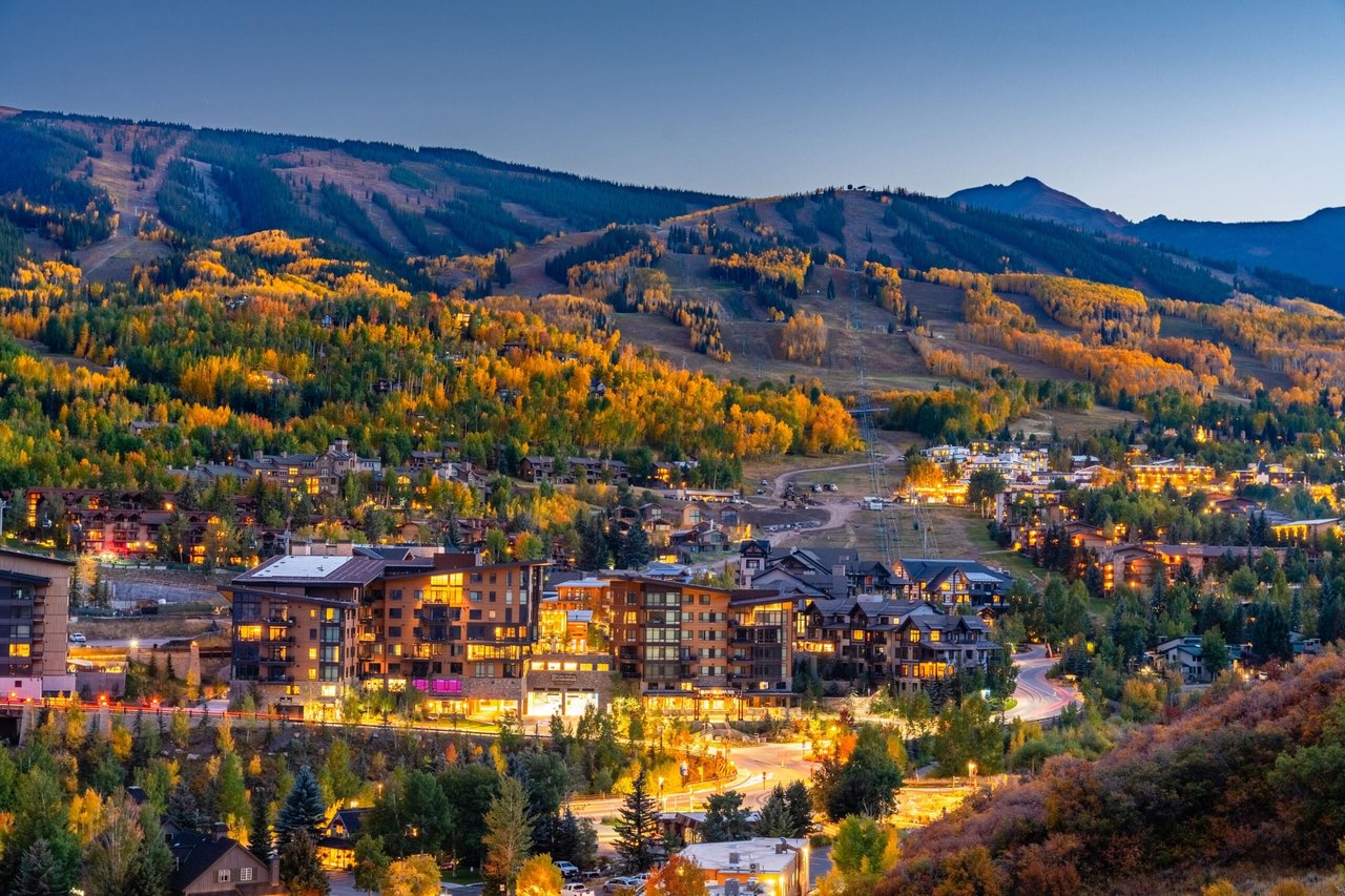 Mountain resort village at dusk, with golden autumn trees and illuminated buildings below the ski slopes.