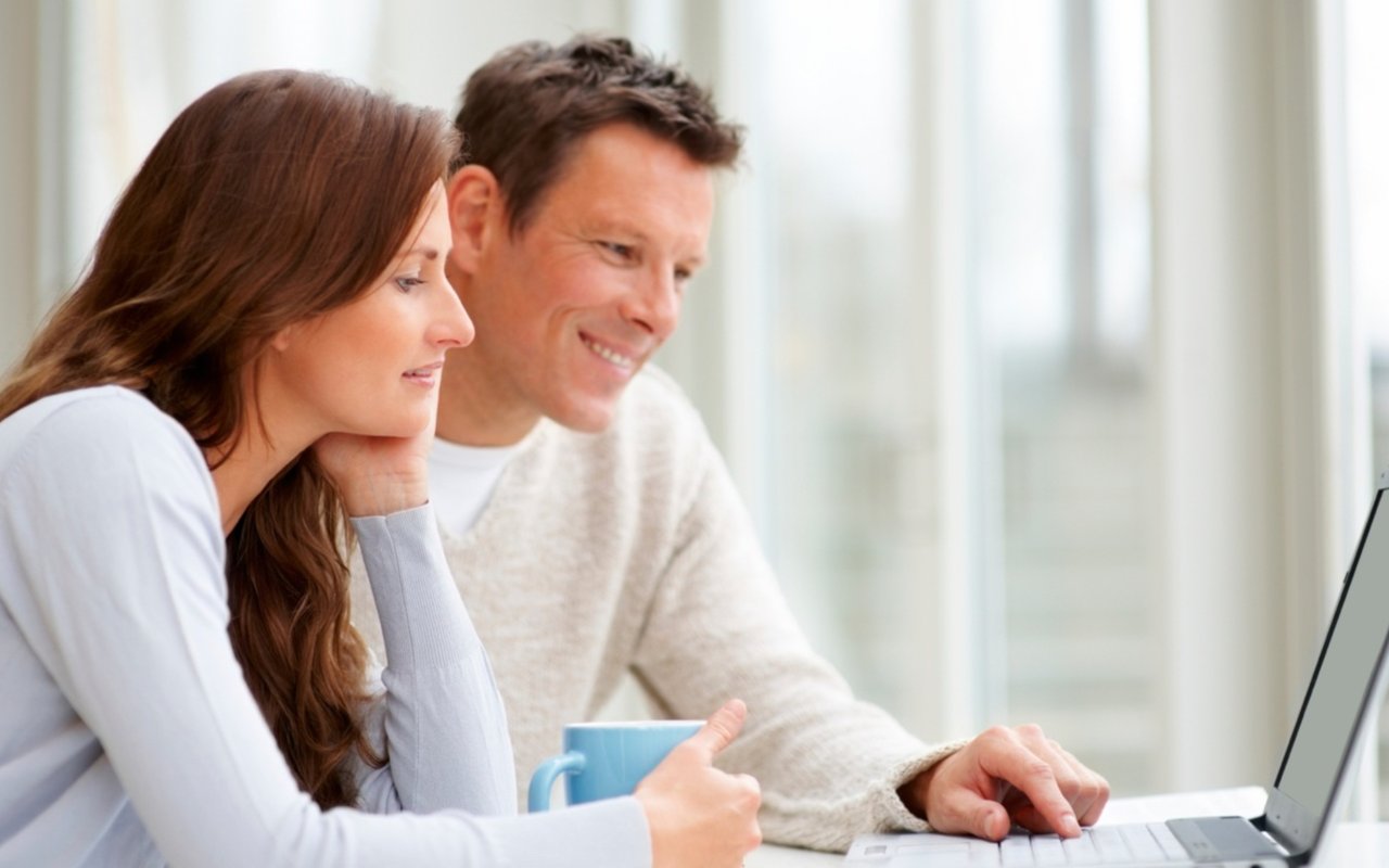 A couple smiles while looking at a laptop together at a table.