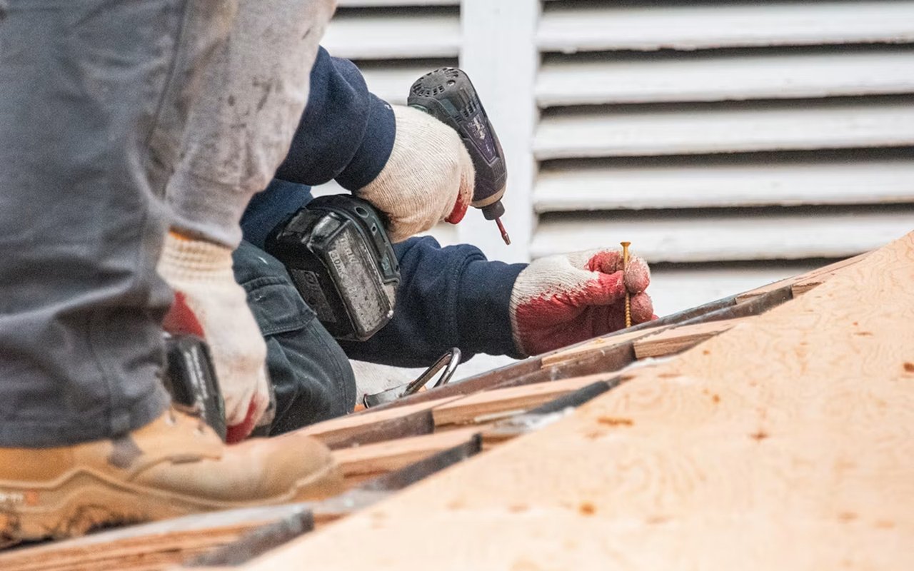 Workers use a drill and hand tools to repair a wooden structure beside white siding.