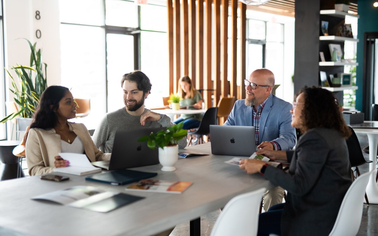Four adults discuss paperwork and laptops around a conference table in a bright modern office.