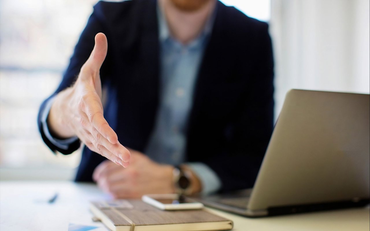 A businessperson extends a hand across a desk beside a laptop and paperwork.