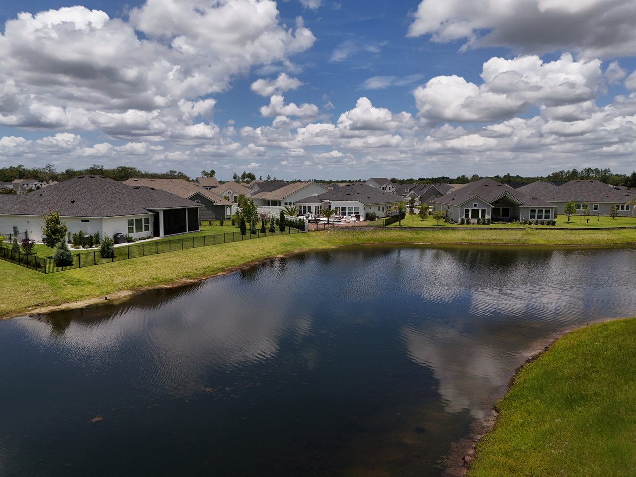 Aerial view of homes along a pond, with fenced lawns and a broad, cloud-filled sky.