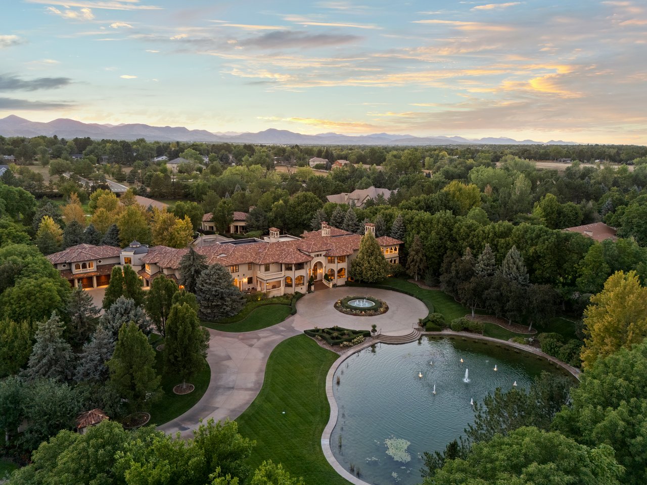 Aerial view of a landscaped estate with a pond, fountain, and mountain ranges at sunset.