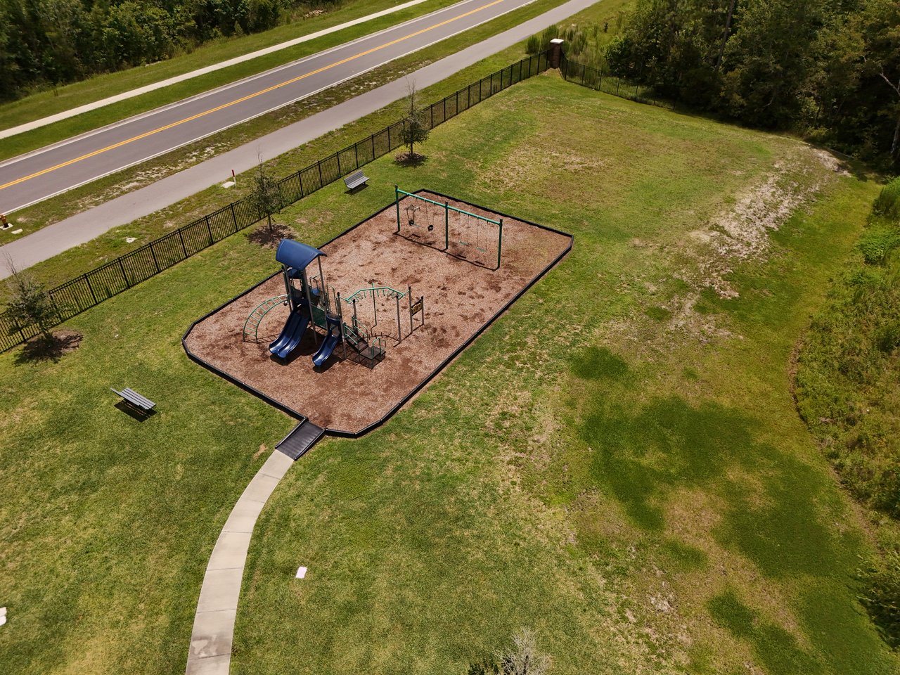 Aerial view of a fenced playground with blue slides and swings amid a grassy open space.