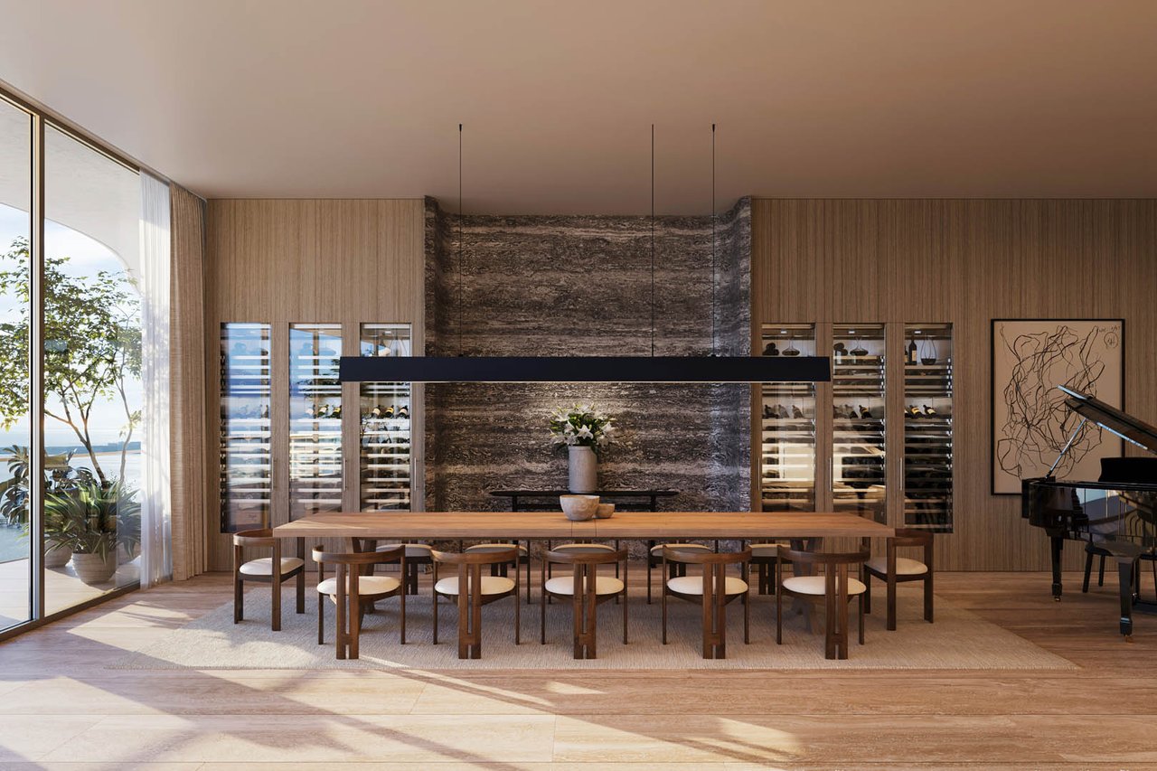 Dining room with a long communal table, wine displays, a grand piano, and water-facing windows.