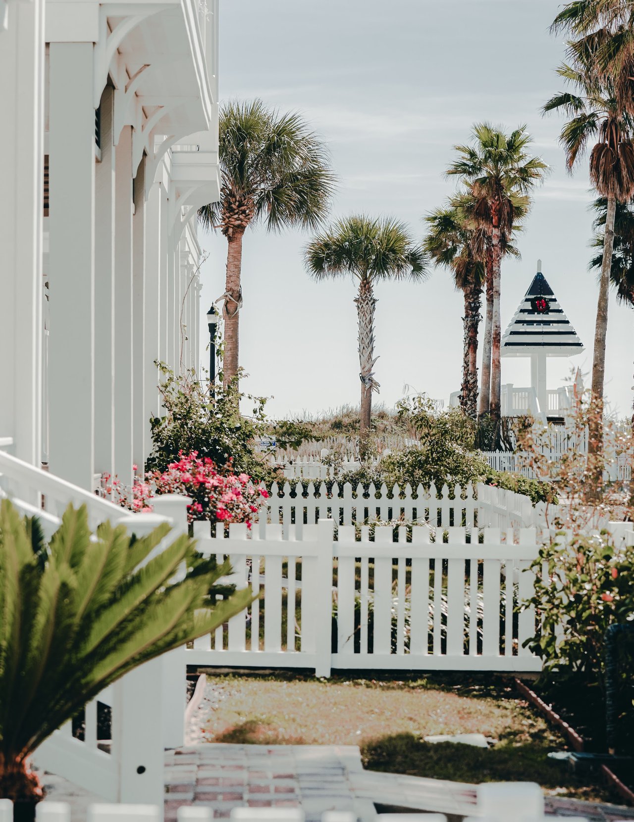 White coastal-style buildings and picket fence among palm trees, with a striped-roof gazebo.