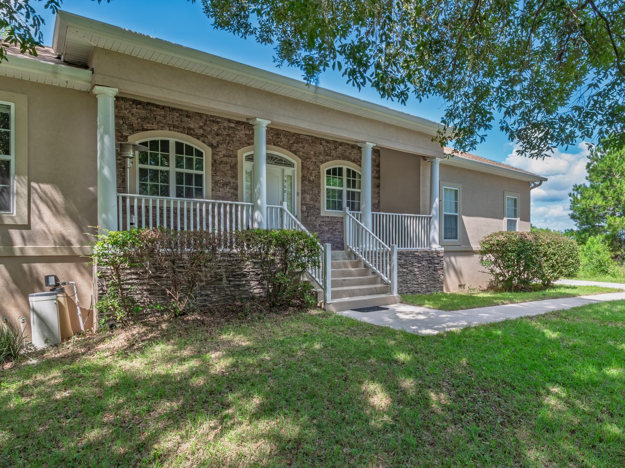 Single-story beige stucco home with a stone-accented entrance, white railings, and a shaded front yard.