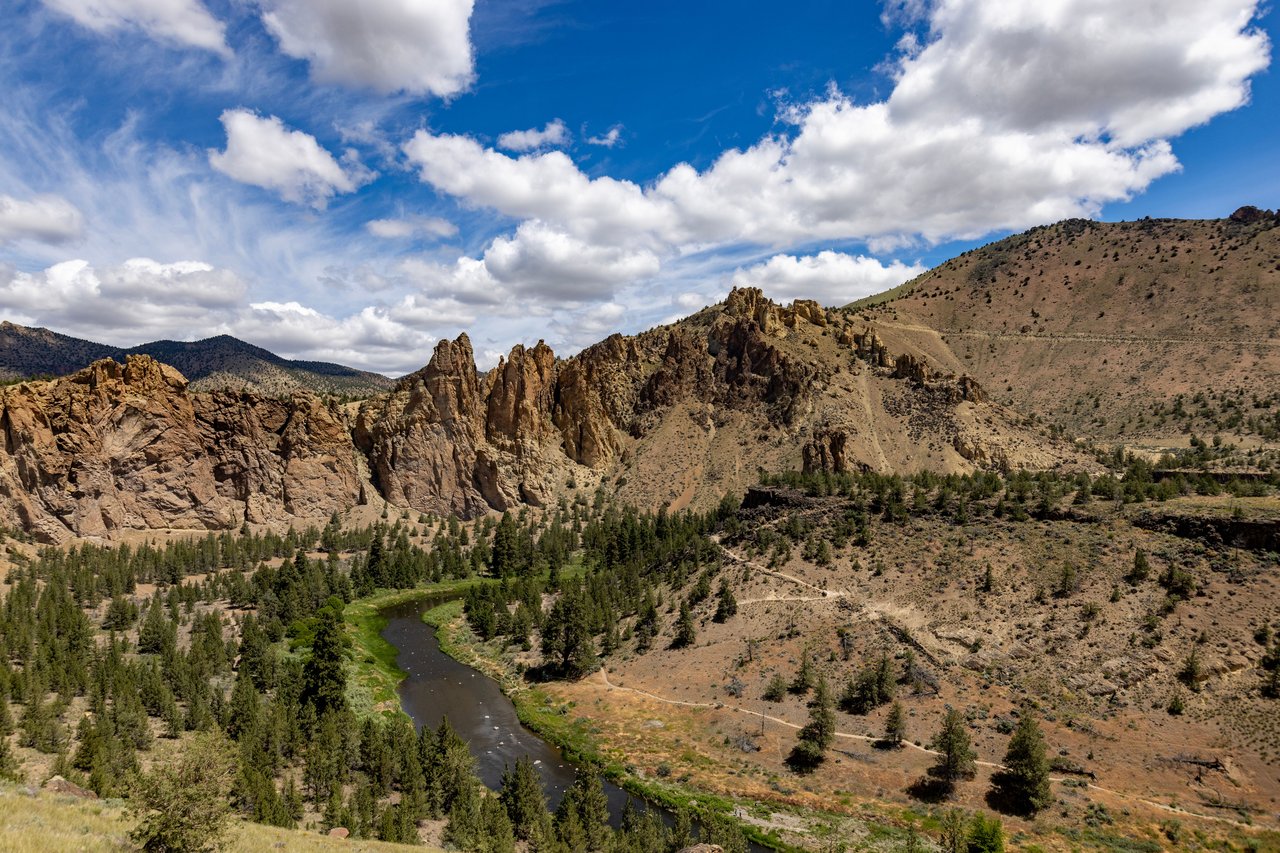 A winding river runs through a rocky canyon dotted with pine trees beneath a cloud-filled sky.