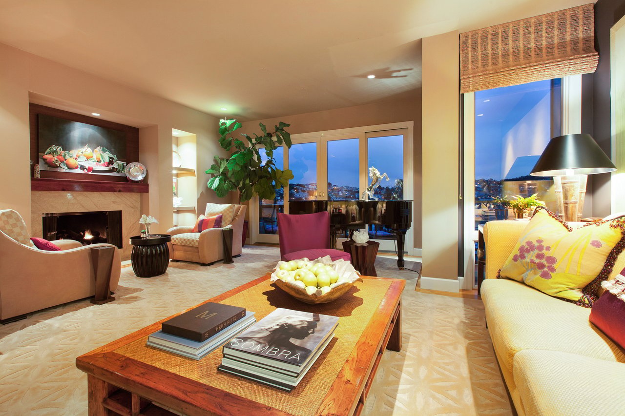 Furnished living room with a fireplace, grand piano, and city views through large windows.