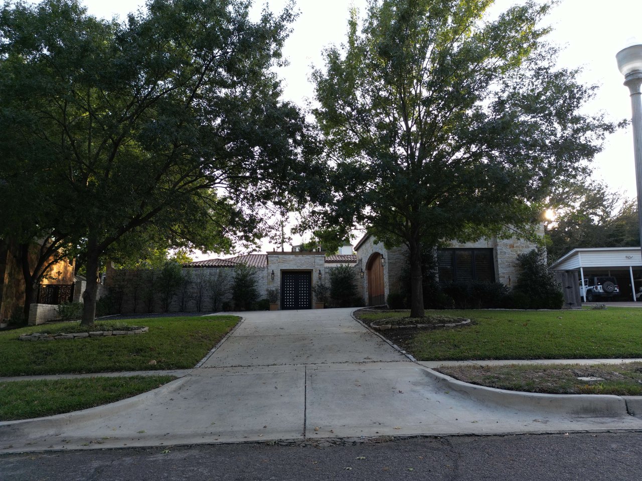 Stone-front home with an arched wooden entrance, broad driveway, and mature shade trees.