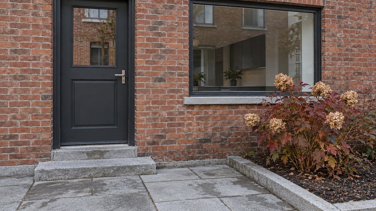 Updated brick residence with a charcoal door, dark-framed window, granite entrance paving, and autumn oakleaf hydrangea.
