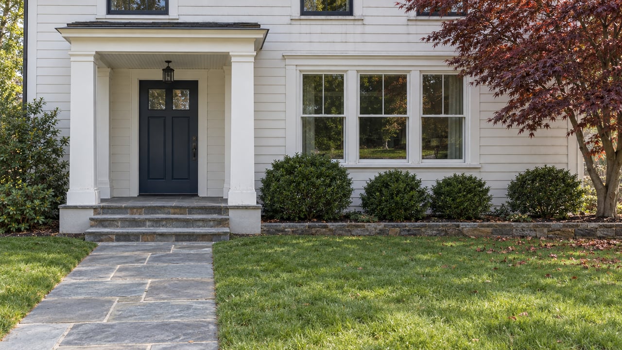 White clapboard home with a covered navy-door entrance, bluestone steps, a broad front lawn and a burgundy Japanese maple.