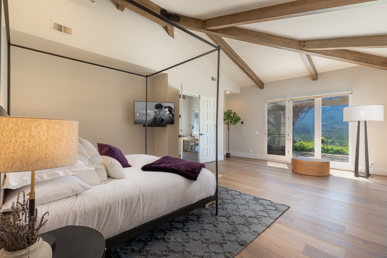 Staged bedroom with a white canopy bed, exposed wood beams, and mountain views through sliding glass doors.