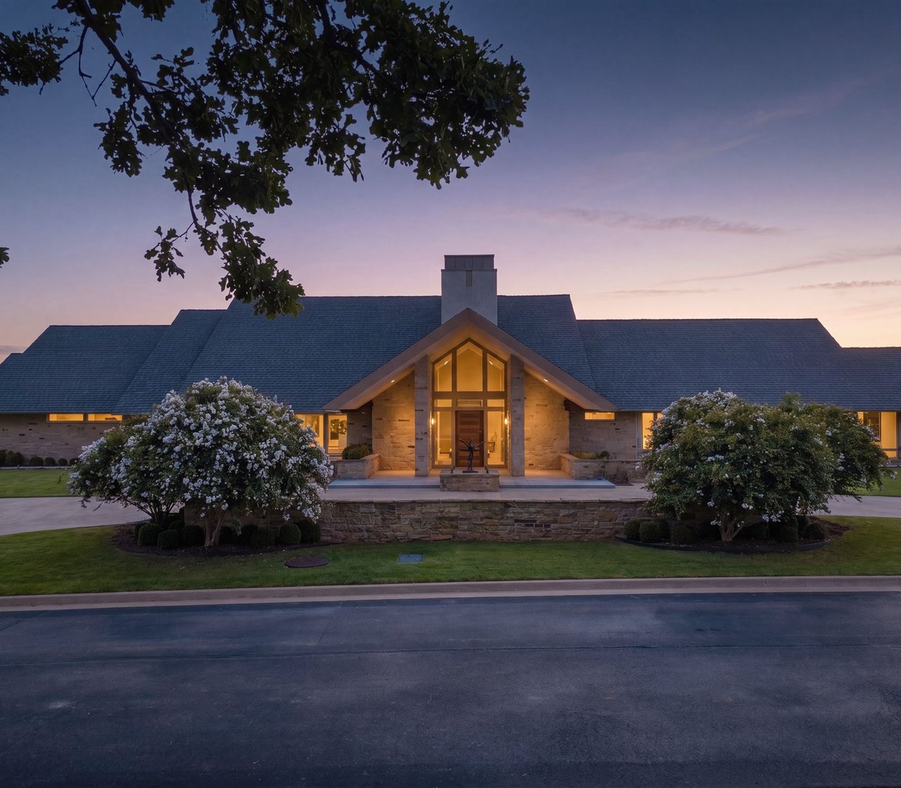 Stone house with a warmly lit entrance and flowering shrubs at dusk.