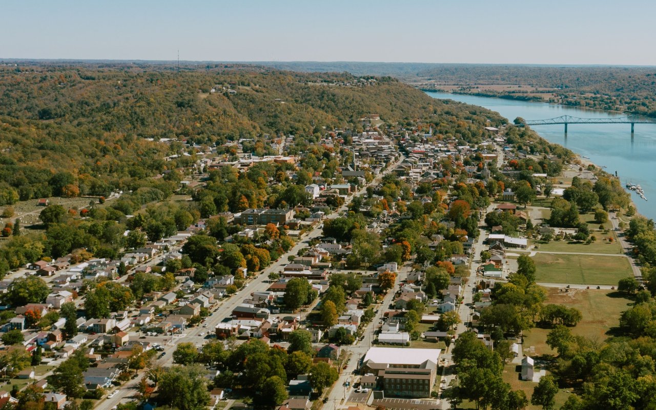 Aerial view of a tree-lined town beside a river, with a bridge in the distance.
