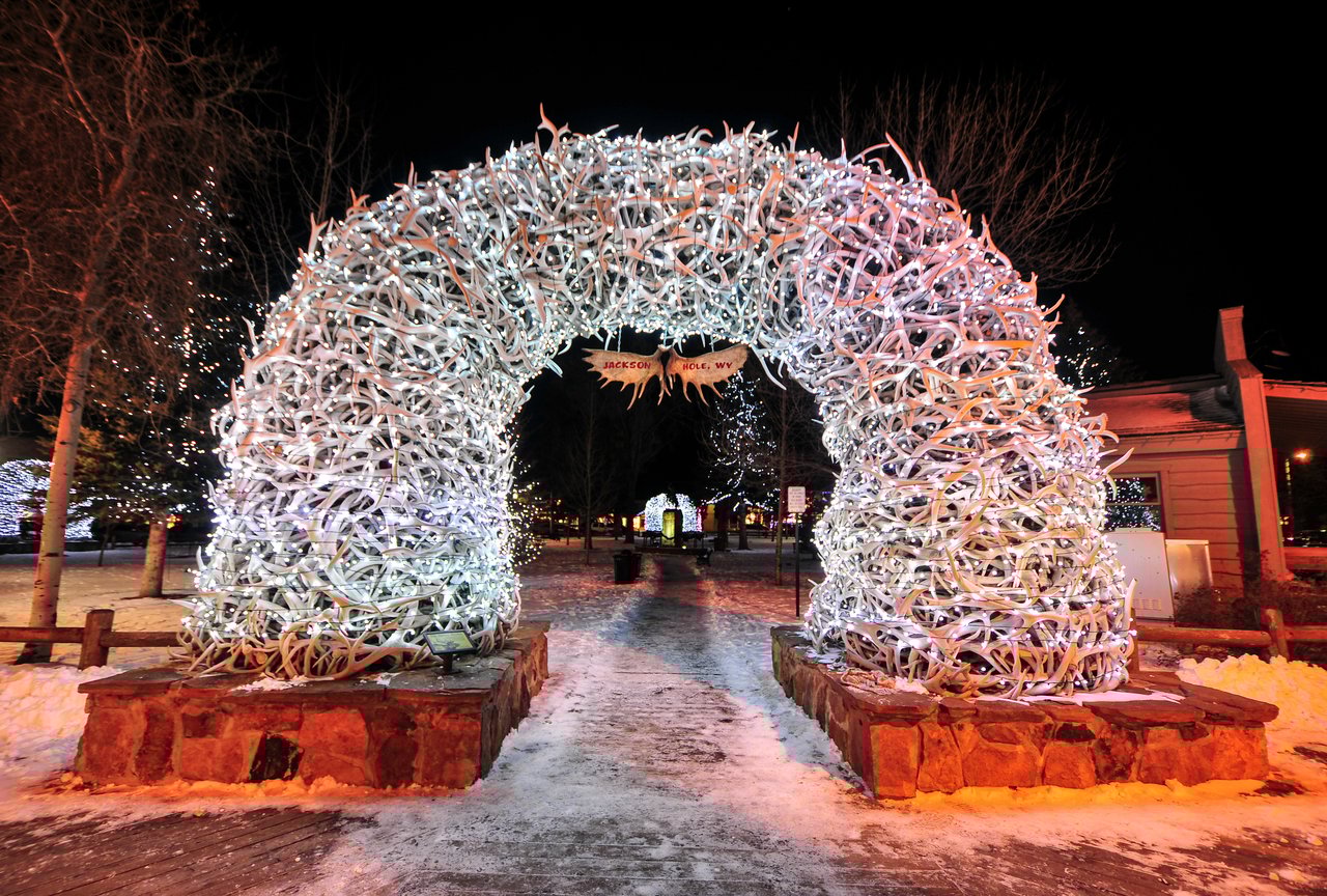 A winter view of Jackson Hole Town Square decorated with holiday lights, featuring snow-covered streets and iconic antler arches, capturing the festive spirit of Jackson, WY.