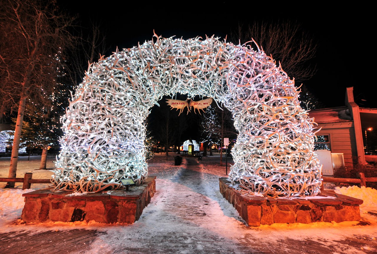 A winter view of Jackson Hole Town Square decorated with holiday lights, featuring snow-covered streets and iconic antler arches, capturing the festive spirit of Jackson, WY.