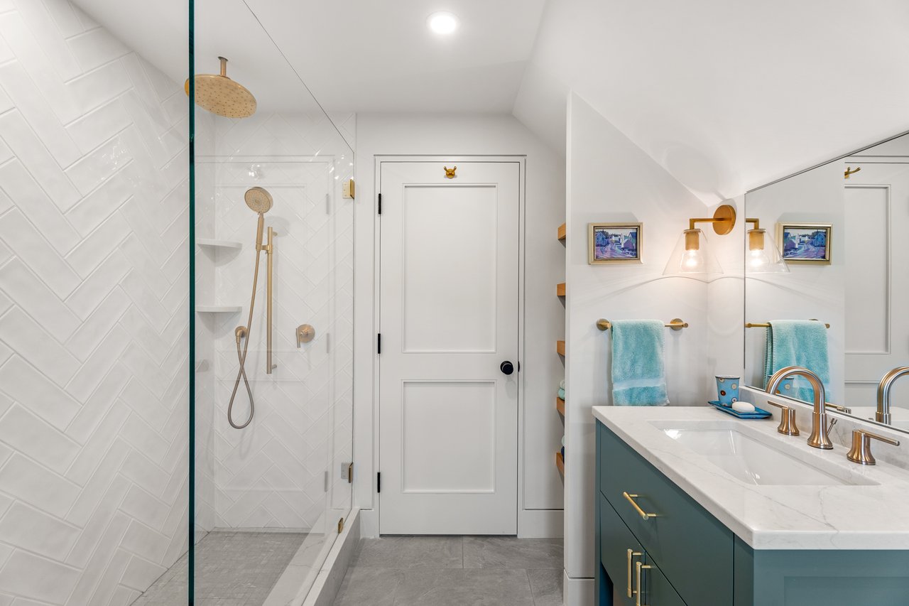 Bathroom with a glass shower, white herringbone tile, teal vanity, and brass fixtures.