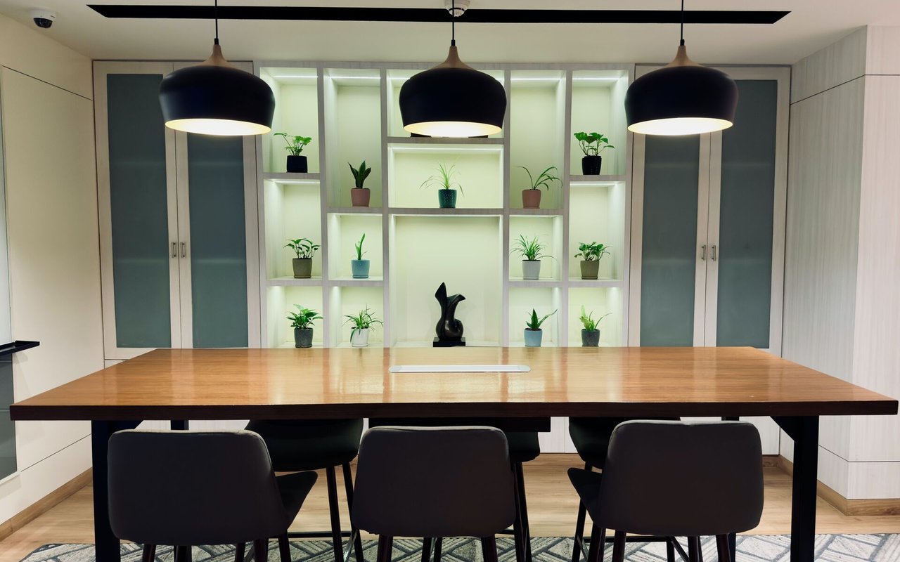Modern meeting room with a long wooden table, pendant lights, and plant-filled illuminated shelving.