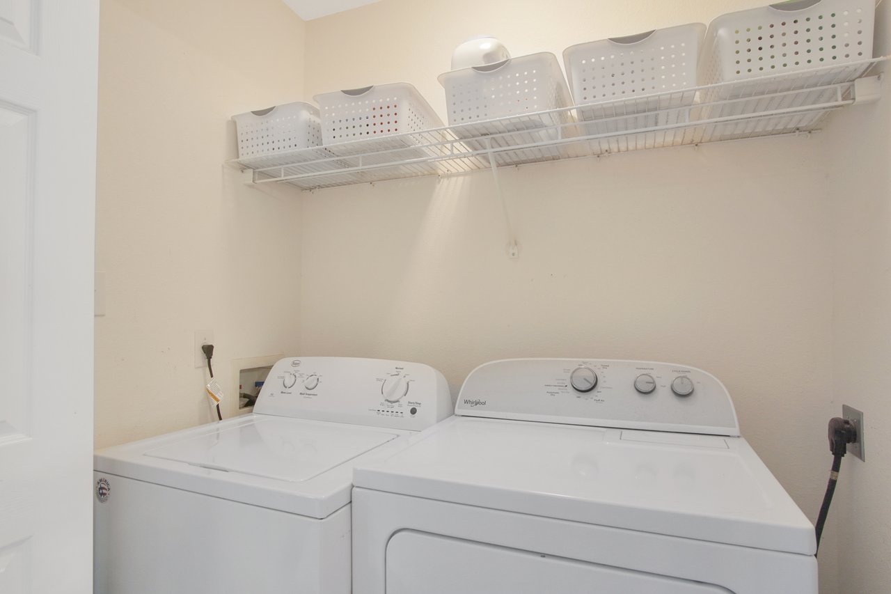 Laundry room with a white washer and dryer side by side, beneath a wire shelf holding white storage baskets. The setting is clean and organized.
