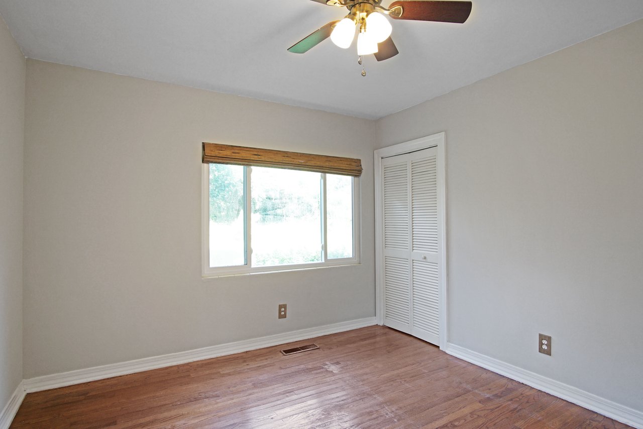 Empty room with wooden floor, beige walls, and a white ceiling fan. A large window with a blind lets in natural light, and there's a white closet door.