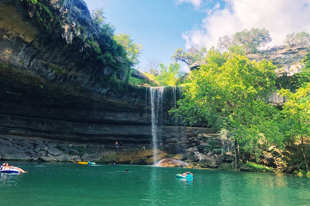 Waterfall over a limestone cliff flowing into a swimming hole with people floating on inner tubes.