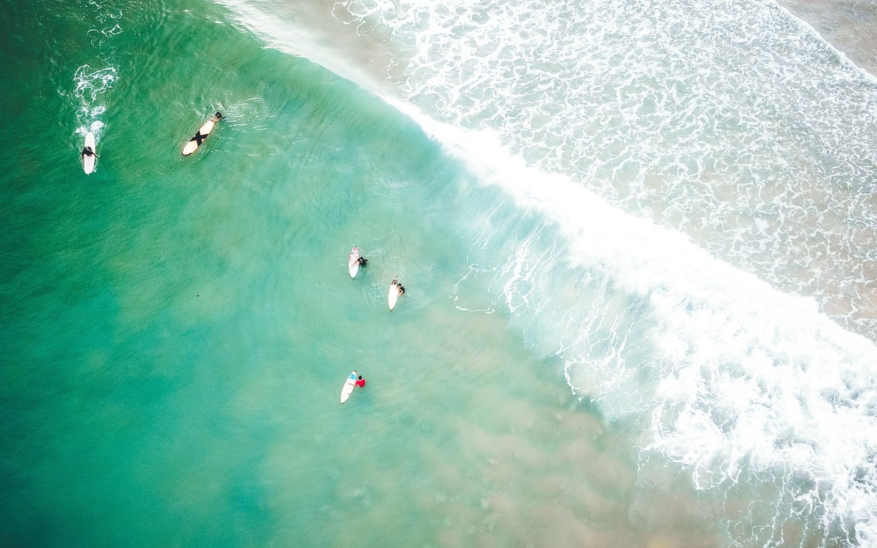Surfers wait in turquoise water as white waves break along the shore.