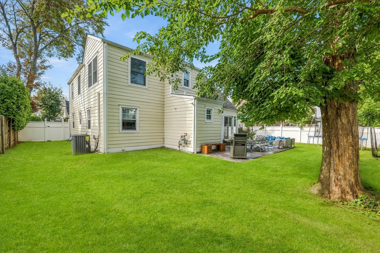 Two-story home behind a broad lawn, with a fenced patio and outdoor seating beneath a large tree.