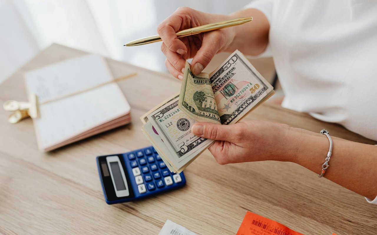 Hands counting U.S. dollar bills beside a calculator and notebook.