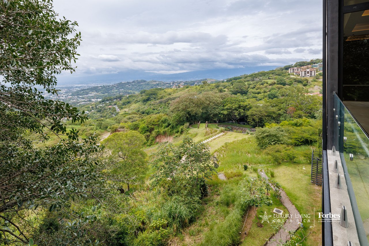 Balcony view over lush hills, landscaped grounds, a distant city, and mountain ridge.