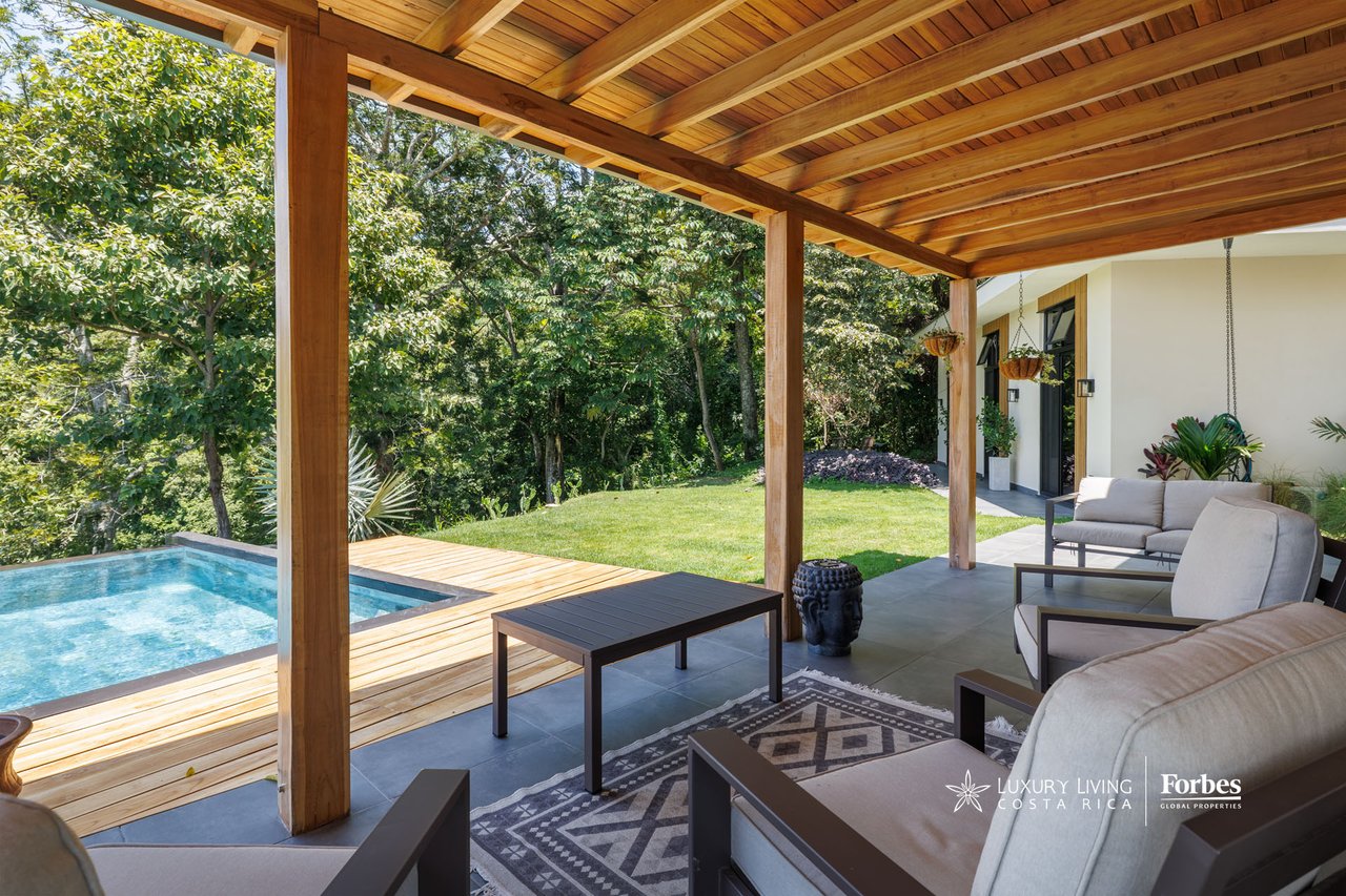 Covered patio with timber ceiling, cushioned seating, and a pool beside a wooded garden.
