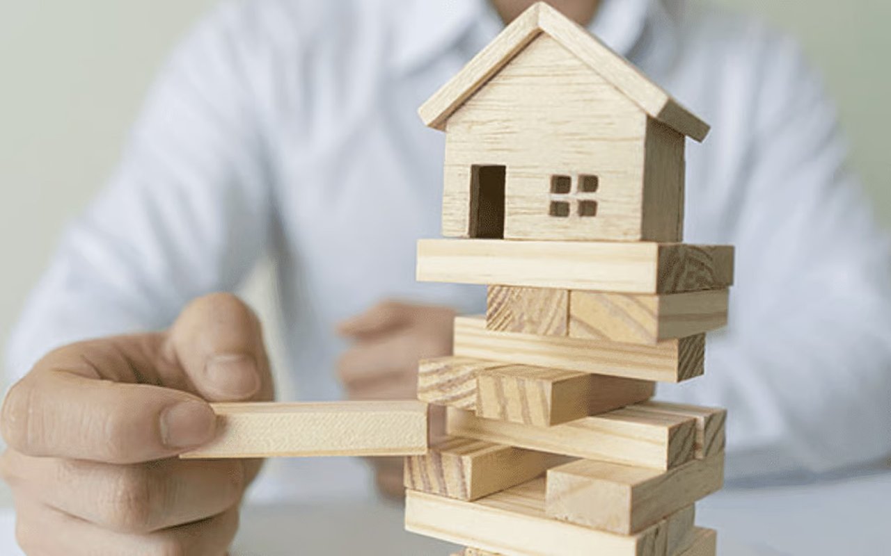 Hand removes a block from a wooden tower topped with a small house.