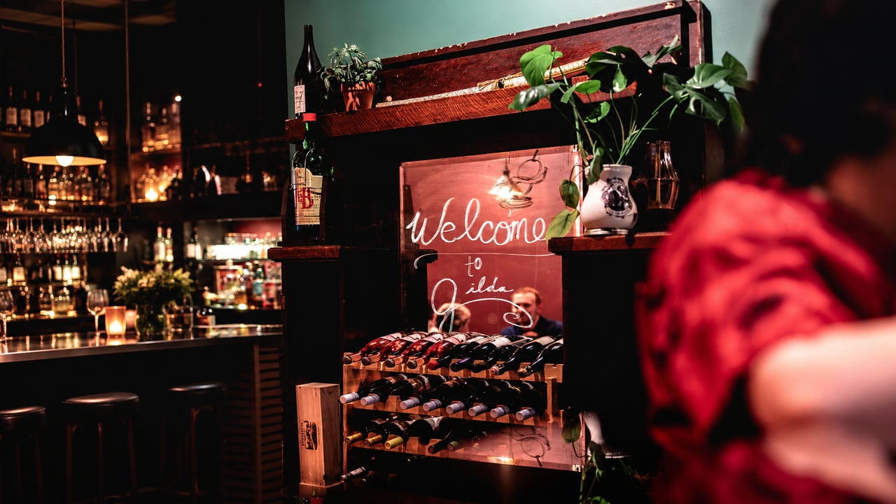 Dimly lit restaurant bar with a wine rack, handwritten welcome sign, and red flowers.