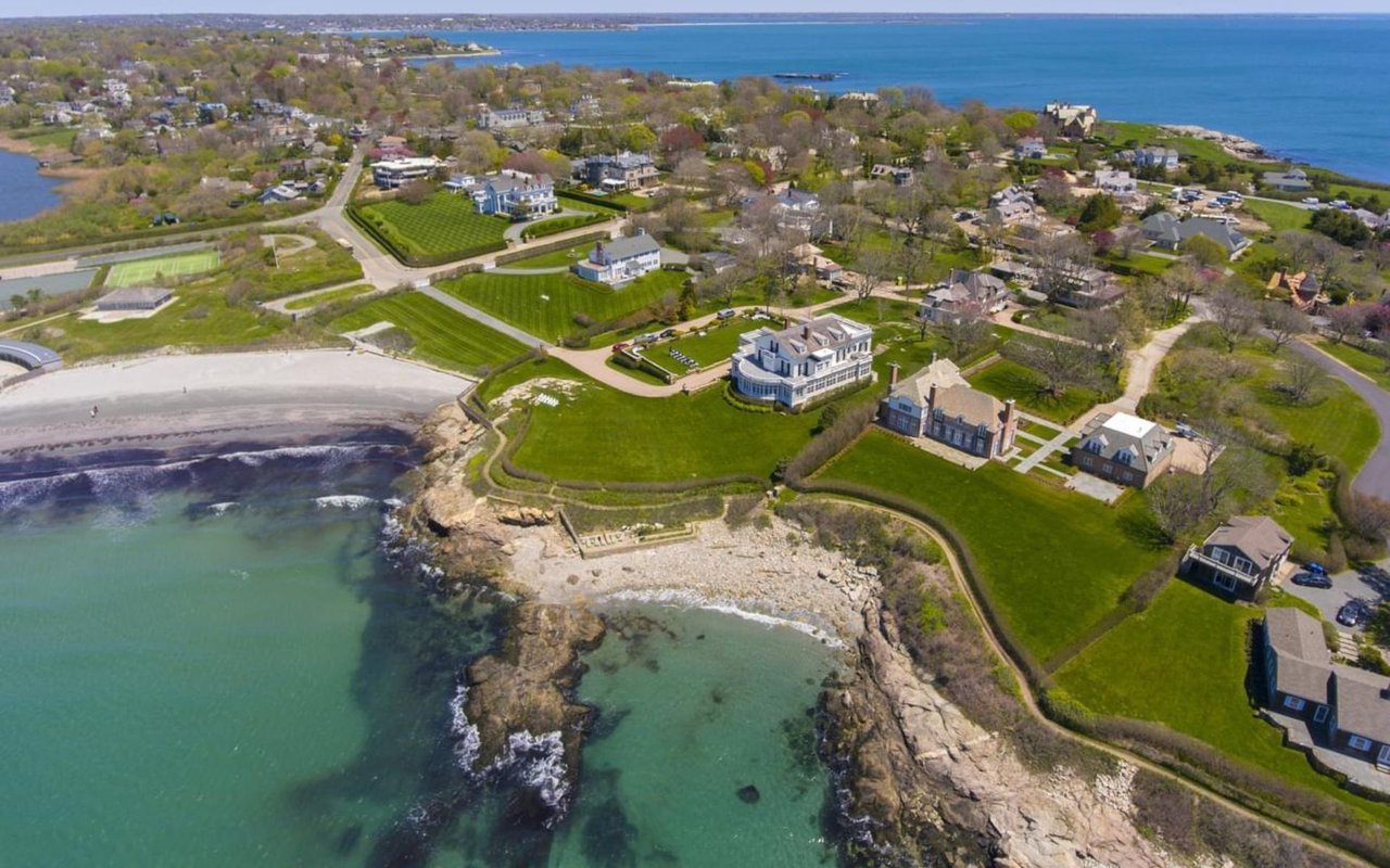 Aerial view of Newport coastal homes, rocky shoreline, sandy beach, and blue-green water.