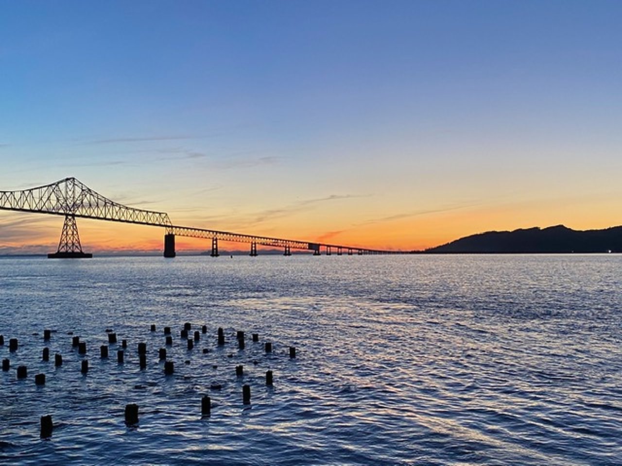 Astoria–Megler Bridge spans the Columbia River at sunset, with wooden pilings in the foreground.