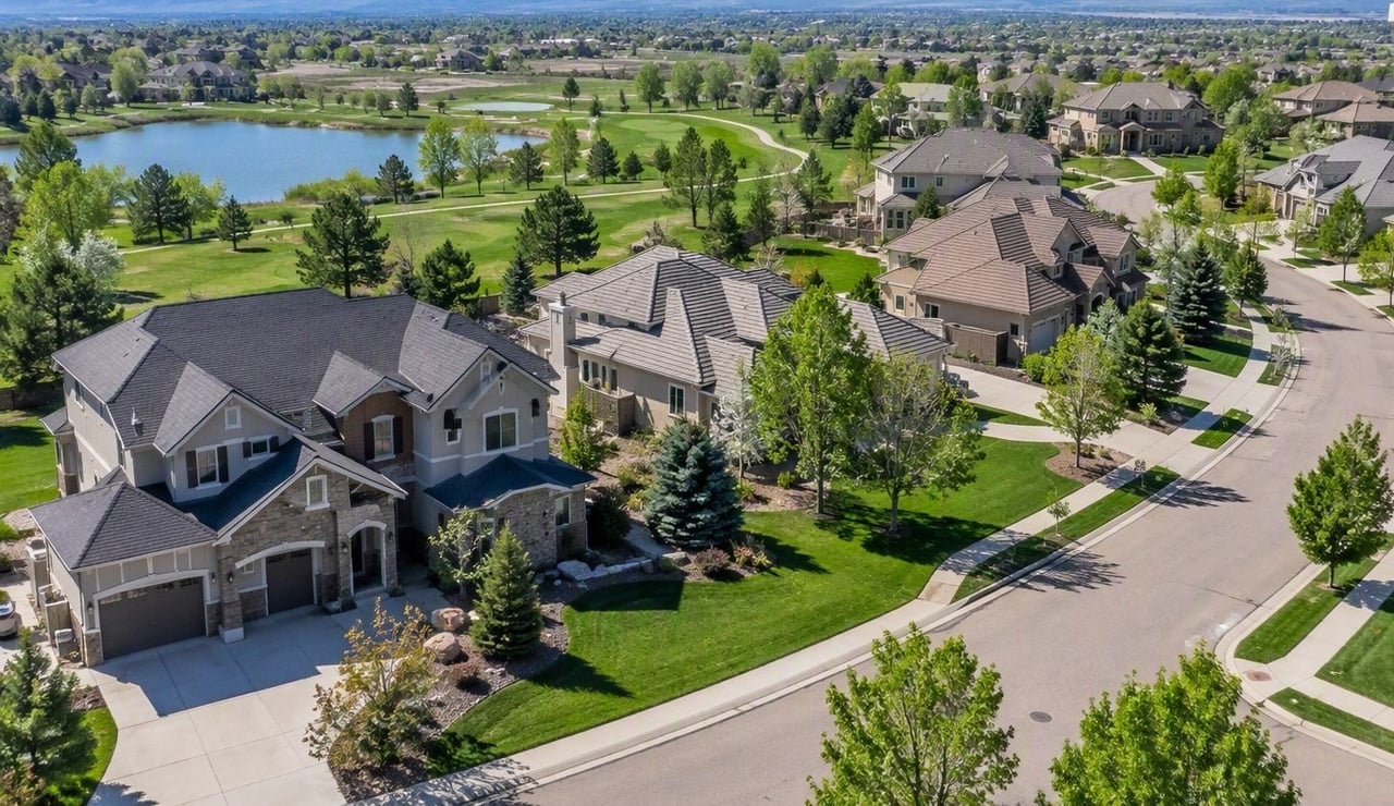Aerial view of large homes beside a lake and park, with a curving street and distant hills.