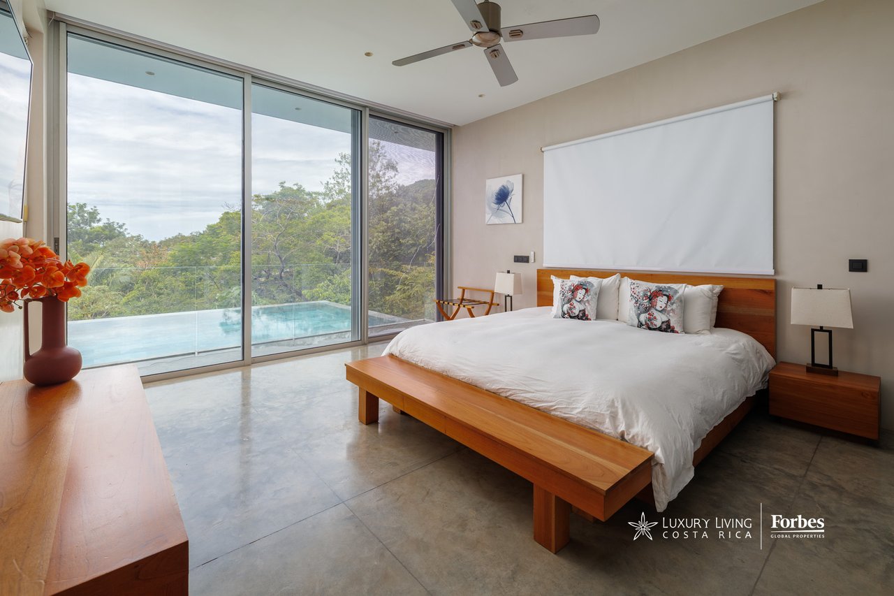 Bedroom with wooden furnishings and sliding glass doors overlooking a pool and green hillside.