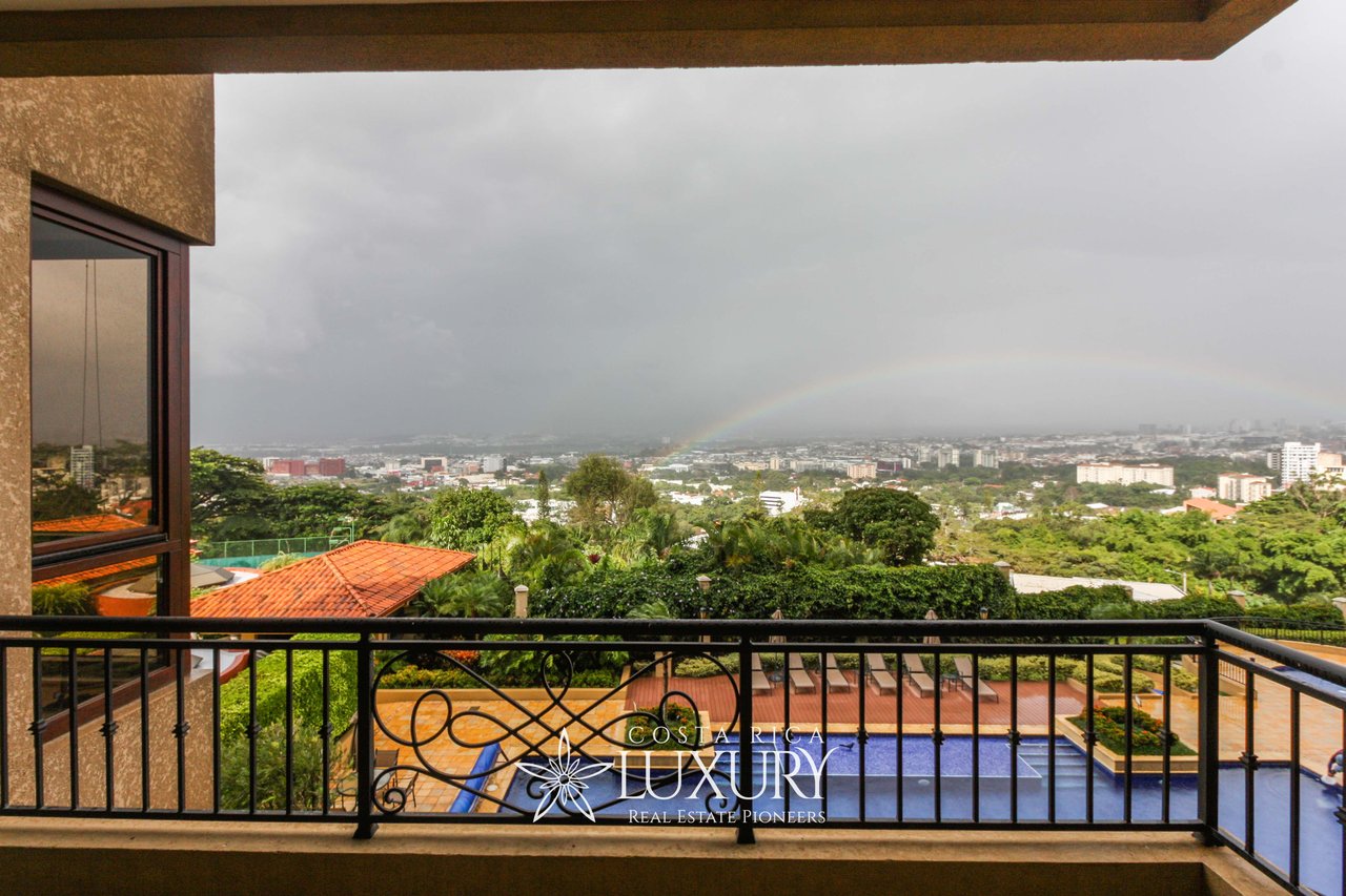Balcony overlooking a swimming pool and green cityscape beneath a rainbow.