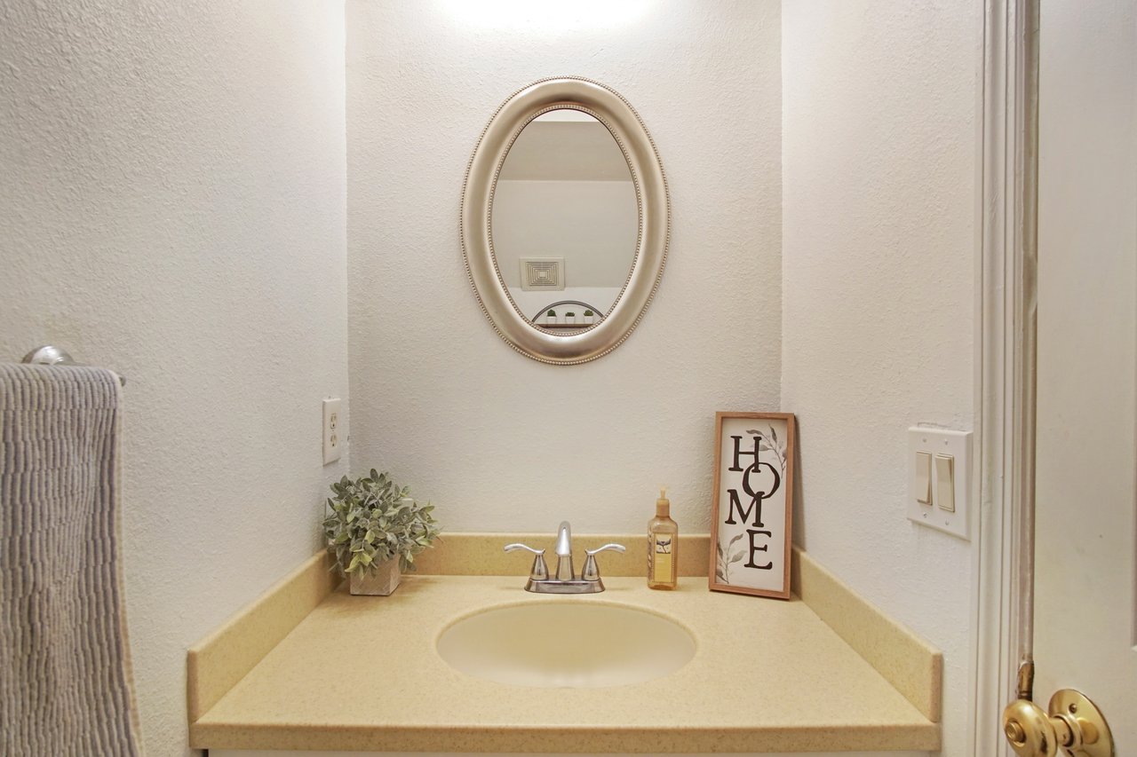 A small bathroom with a beige countertop and oval mirror. A potted plant, hand soap, and a framed "HOME" sign are on the counter, creating a cozy feel.