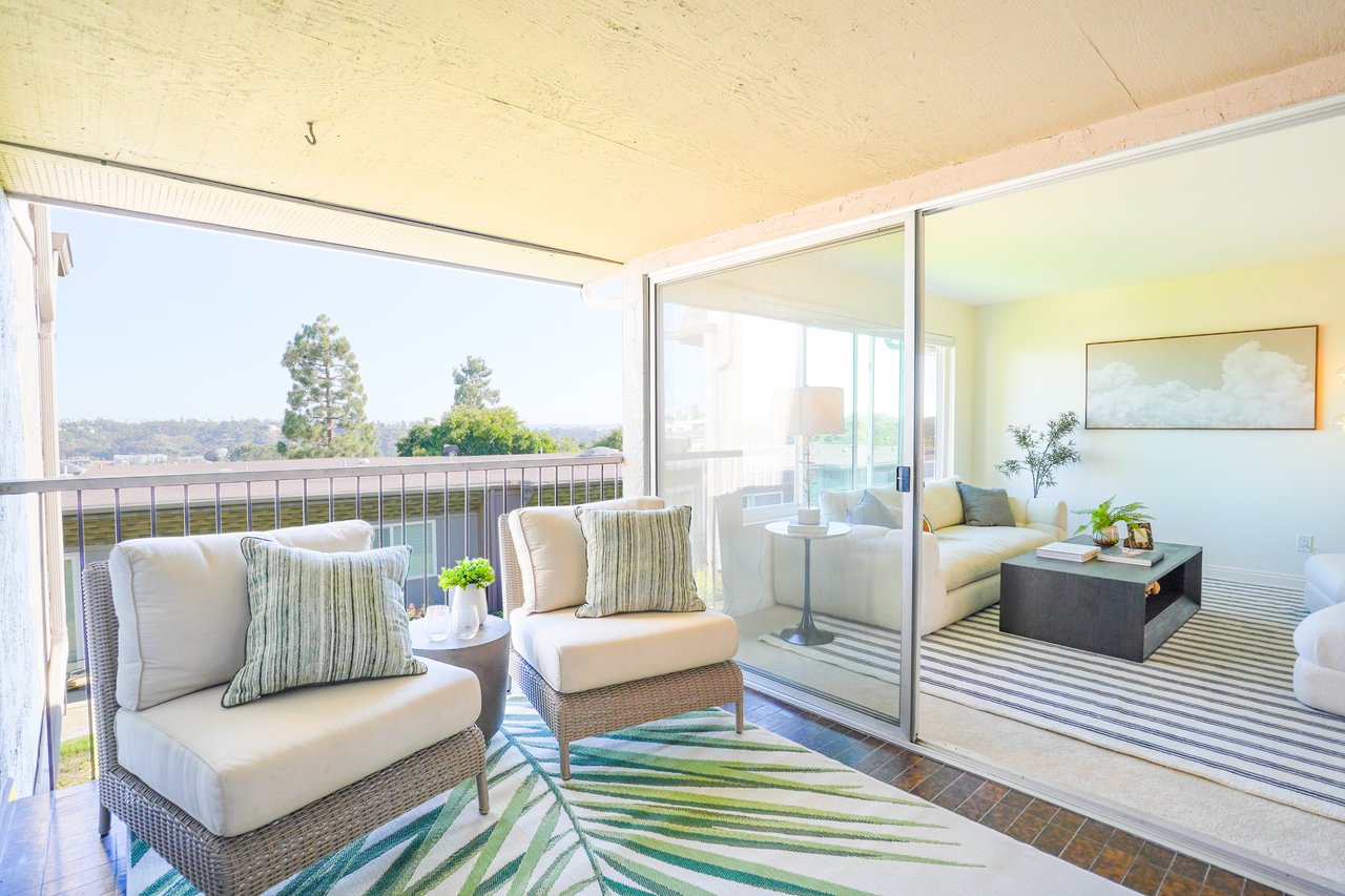 Inviting private balcony at 6321 Caminito Luisito, San Diego CA 92111, featuring outdoor seating with cushions, green accent rug, and sliding glass doors opening to a bright modern living room with natural light and hillside views.