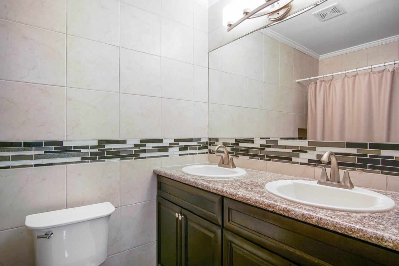 Elegant bathroom with double sinks on a granite countertop, warm beige tiles, and a subtle modern mosaic border. Soft lighting creates a cozy ambiance.