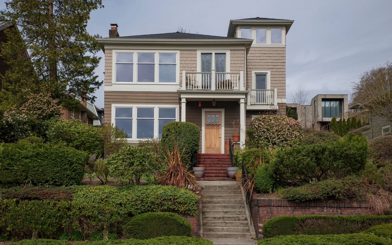 Two-story taupe house with white trim, a central staircase, and landscaped front garden.