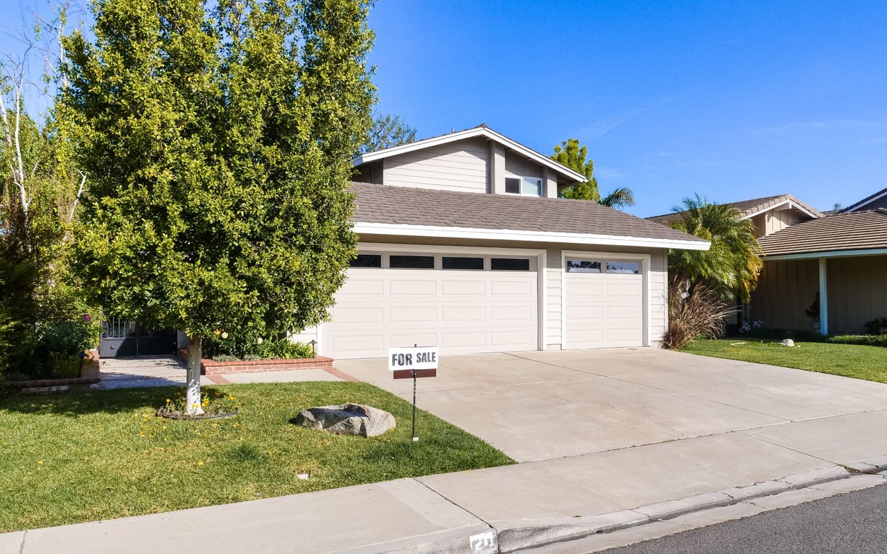 Two-story home with a wide garage, leafy front yard, and a FOR SALE sign.