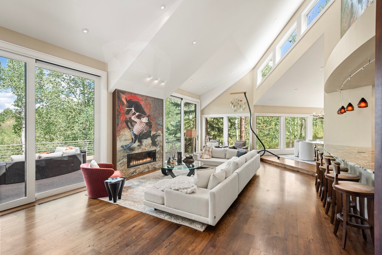 Living room with vaulted ceiling, expansive windows, a sculptural fireplace, and an open kitchen.
