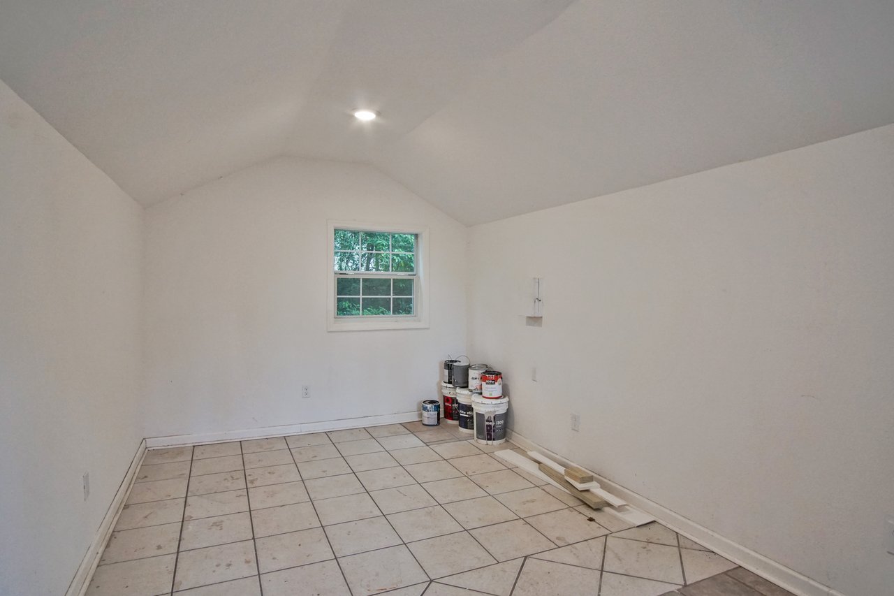 Minimalist, small room with white walls, tiled floor, and a single window. Paint buckets and wood planks are stacked in the corner.