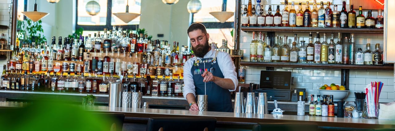 Bartender mixing a drink behind a bottle-lined bar with pendant lights and tiled walls.