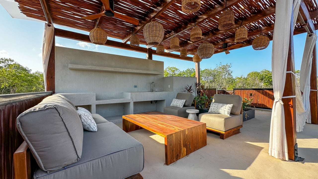 Outdoor lounge with gray seating, concrete kitchen, and woven lanterns beneath a timber pergola.