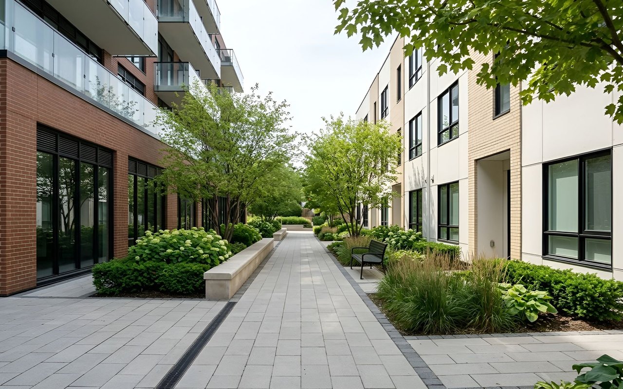 Landscaped courtyard walkway between modern condominium buildings, with planting beds and a bench.